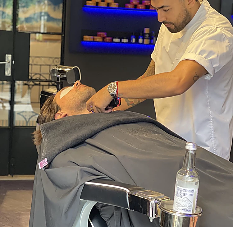 Barbero de camisa blanca afeitando a un cliente recostado con los ojos cubiertos, junto a las repisas con luz azul