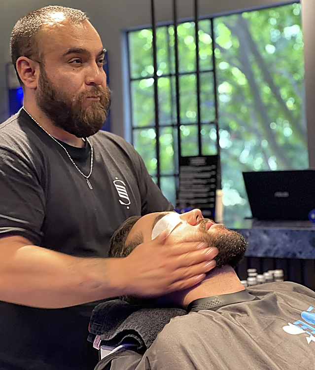 Barbero aplicando un tratamiento facial a un cliente recostado con los ojos cubiertos, frente al ventanal