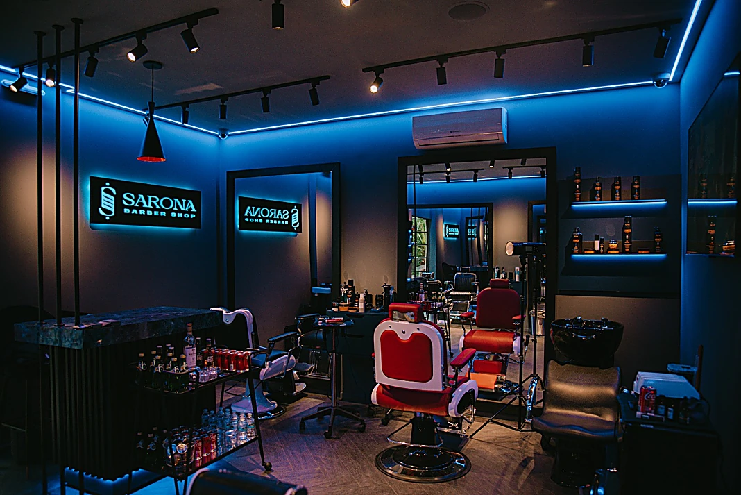 Interior de Sarona: el letrero SARONA Barber Shop iluminado en la pared marino, un sillón de barbero rojo con base cromada y la línea de luz azul en el techo