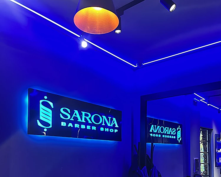 Letrero SARONA Barber Shop iluminado en cian sobre la pared azul, con la lámpara colgante naranja encima