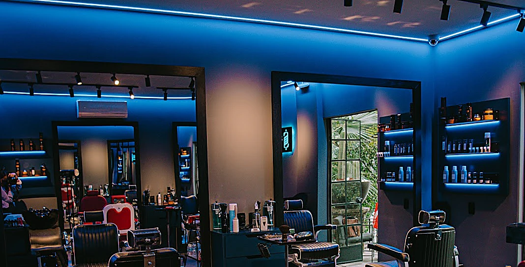 El salón completo de Sarona: sillones de barbero, espejos con marco negro, repisas y la línea de luz azul del techo