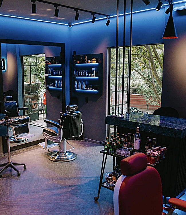 Sillones de barbero junto a un ventanal con árboles, espejos y repisas iluminadas en azul